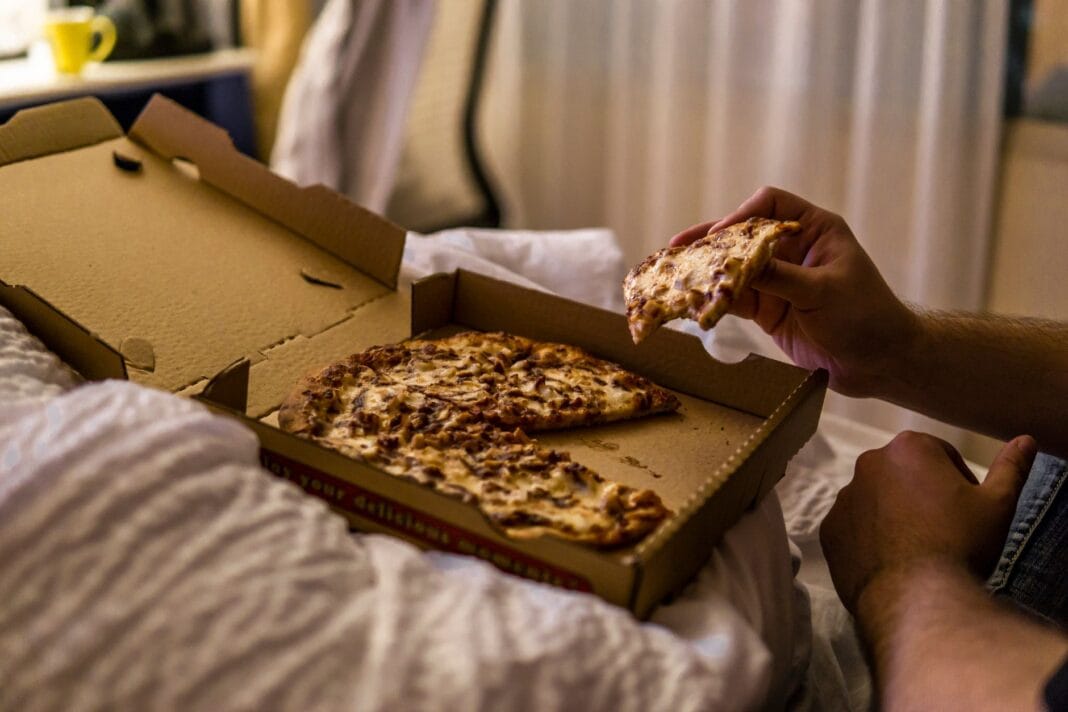 Mężczyzna je kawałek pizzy z pudełka w łóżku wieczorem Mężczyzna je kawałek pizzy z pudełka w łóżku wieczorem
