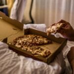 Mężczyzna je kawałek pizzy z pudełka w łóżku wieczorem